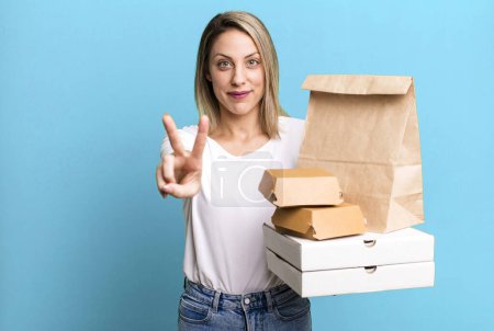 Foto de Pretty blonde woman smiling and looking friendly, showing number two. paper fast food take away packages - Imagen libre de derechos