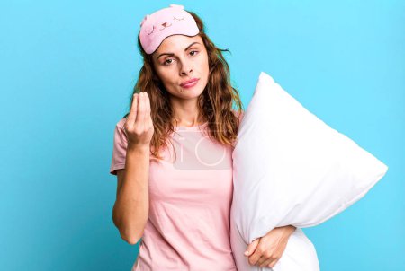 Photo for Hispanic pretty woman making capice or money gesture, telling you to pay wearing pajamas and a pillow - Royalty Free Image