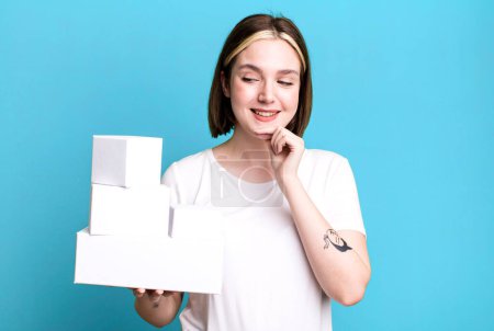 Photo for Young pretty woman smiling with a happy, confident expression with hand on chin. white blank boxes - Royalty Free Image