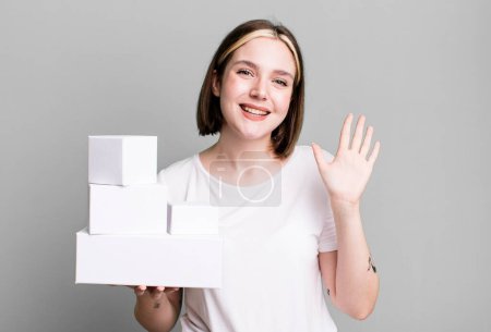 Photo for Young pretty woman smiling happily, waving hand, welcoming and greeting you. white blank boxes - Royalty Free Image