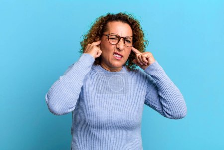 Téléchargez les photos : Femme hispanique d'âge moyen regardant en colère, stressé et agacé, couvrant les deux oreilles à un bruit assourdissant, son ou musique forte - en image libre de droit