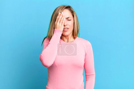 Foto de Blonde adult woman looking sleepy, bored and yawning, with a headache and one hand covering half the face - Imagen libre de derechos