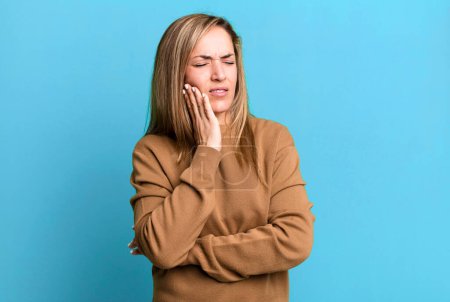 Blonde erwachsene Frau hält Wange und leidet unter schmerzhaften Zahnschmerzen, fühlt sich krank, elend und unglücklich, sucht einen Zahnarzt
