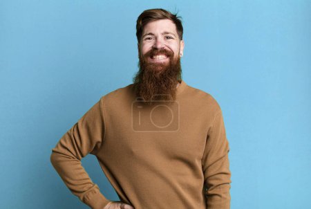 Foto de Barba larga y hombre de pelo rojo sonriendo felizmente con una mano en la cadera y confiado - Imagen libre de derechos