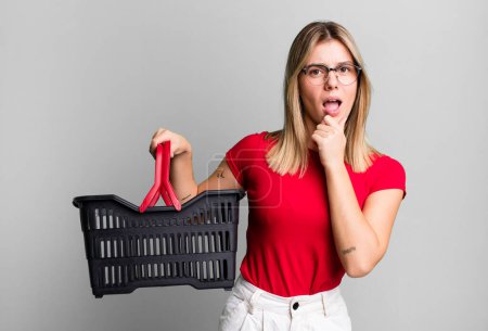 Foto de Young pretty woman with mouth and eyes wide open and hand on chin. empty shopping basket concept - Imagen libre de derechos