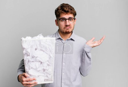 Téléchargez les photos : Jeune homme caucasien adulte se sentant perplexe et confus et doutant avec un concept de poubelle boules de papier - en image libre de droit