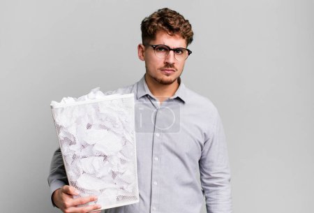 Téléchargez les photos : Jeune homme caucasien adulte se sentant triste, contrarié ou en colère et regardant vers le côté avec un concept de poubelle boules de papier - en image libre de droit