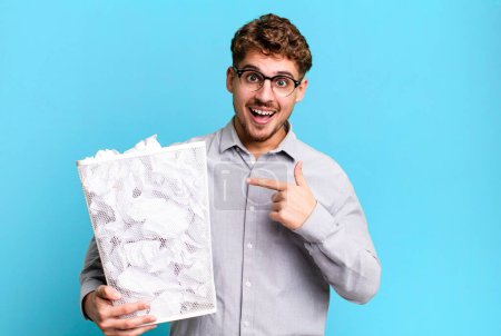 Photo for Young adult caucasian man looking excited and surprised pointing to the side with a paper balls trash concept - Royalty Free Image
