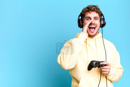Téléchargez les photos : Young adult caucasian man feeling happy,giving a big shout out with hands next to mouth with headset and a controller. gamer concept - en image libre de droit