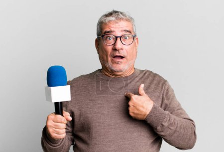 Photo for Middle age senior man looking shocked and surprised with mouth wide open, pointing to self. journalist or tv presenter with a micro - Royalty Free Image
