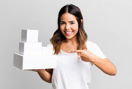 Téléchargez les photos : Hispanique jolie femme souriant joyeusement, se sentant heureux et pointant vers le côté avec des boîtes de paquets vides - en image libre de droit
