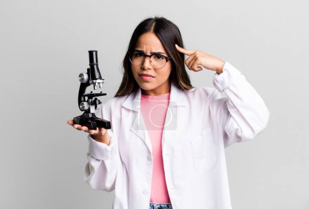 Photo for Hispanic pretty woman feeling confused and puzzled, showing you are insane. scients student with a microscope - Royalty Free Image