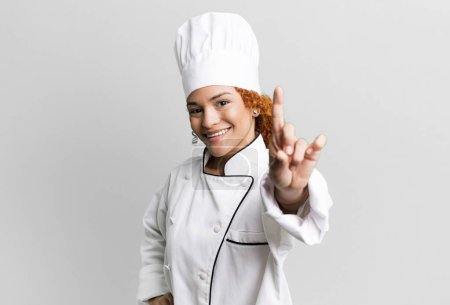 Foto de Cabello rojo bonita mujer sonriendo orgullosa y confiadamente haciendo número uno. concepto de chef - Imagen libre de derechos