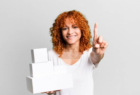 Photo for Red hair pretty woman smiling and looking friendly, showing number one. blank packages boxes concept - Royalty Free Image