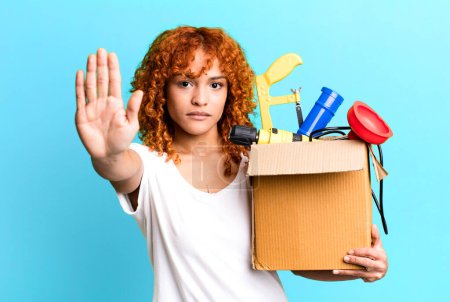 Photo for Red hair pretty woman looking serious showing open palm making stop gesture. housekeeper and toolbox concept - Royalty Free Image