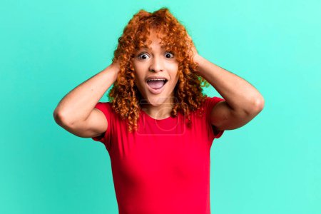 Photo for Redhair pretty woman raising hands to head, open-mouthed, feeling extremely lucky, surprised, excited and happy - Royalty Free Image