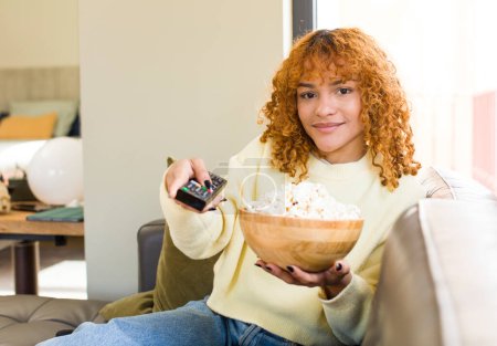 Téléchargez les photos : Jeune rousse cheveux latins jolie femme manger popcorns sur un canapé et regarder un film - en image libre de droit
