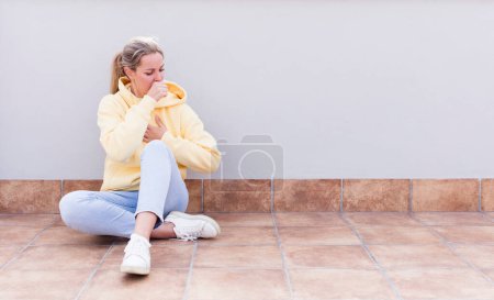 Téléchargez les photos : Jolie femme assise caucasienne se sentant malade avec un mal de gorge et des symptômes de grippe, toux avec la bouche couverte - en image libre de droit