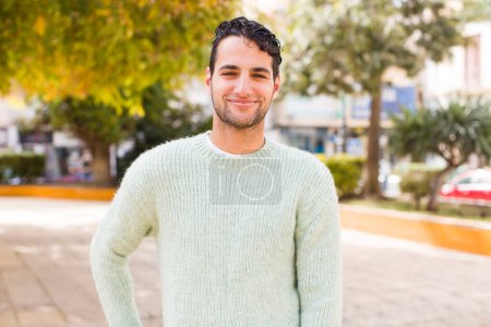 Photo for Young hispanic man smiling happily with a hand on hip and confident, positive, proud and friendly attitude - Royalty Free Image
