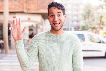 Téléchargez les photos : Jeune homme hispanique souriant joyeusement et joyeusement, agitant la main, vous accueillant et vous saluant, ou disant au revoir - en image libre de droit