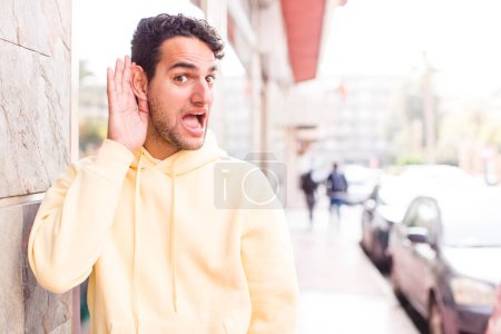 Téléchargez les photos : Jeune homme hispanique regardant heureux, écoutant, essayant d'entendre une conversation secrète ou des commérages, écoutant - en image libre de droit