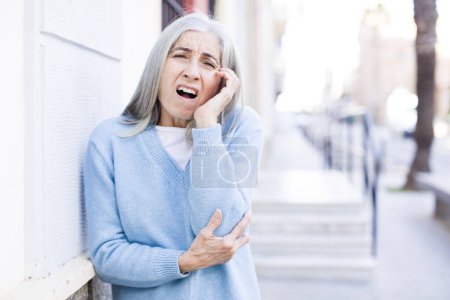 Photo for Senior retired pretty white hair woman open-mouthed in shock and disbelief, with hand on cheek and arm crossed, feeling stupefied and amazed - Royalty Free Image