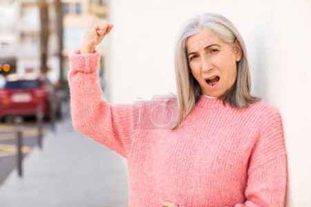 Photo for Senior retired pretty white hair woman feeling happy, satisfied and powerful, flexing fit and muscular biceps, looking strong after the gym - Royalty Free Image