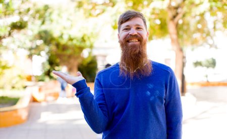 Téléchargez les photos : Homme barbu de cheveux roux souriant, se sentant confiant, réussi et heureux, montrant concept ou idée sur l'espace de copie sur le côté - en image libre de droit