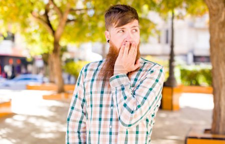 Photo for Red hair bearded man feeling shocked and astonished holding face to hand in disbelief with mouth wide open - Royalty Free Image