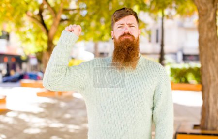 Téléchargez les photos : Cheveux roux barbu homme se sentant sérieux, fort et rebelle, levant le poing, protestant ou se battant pour la révolution - en image libre de droit