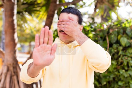 Photo for Young cool man covering face with hand and putting other hand up front to stop camera, refusing photos or pictures - Royalty Free Image