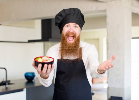 Téléchargez les photos : Homme aux cheveux roux souriant heureux et offrant ou montrant un concept avec un bol de ramen. concept de chef - en image libre de droit