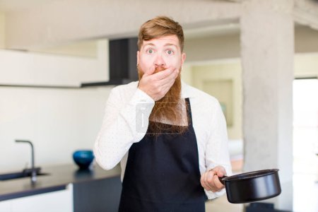 Foto de Hombre de pelo rojo que cubre la boca con una mano y la expresión impactada o sorprendida en una cocina. concepto de chef - Imagen libre de derechos