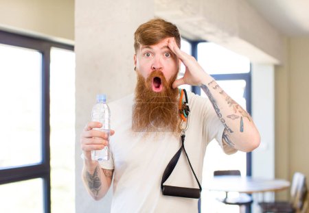 Téléchargez les photos : Homme aux cheveux roux regardant heureux, étonné et surpris avec une bouteille d'eau. concept de fitness - en image libre de droit