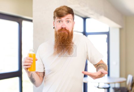 Téléchargez les photos : Homme aux cheveux roux haussant les épaules, se sentant confus et incertain avec une bouteille de jus d'orange - en image libre de droit