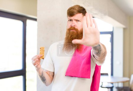 Photo for Red hair man looking serious showing open palm making stop gesture with a cereal bar. fitness concept - Royalty Free Image