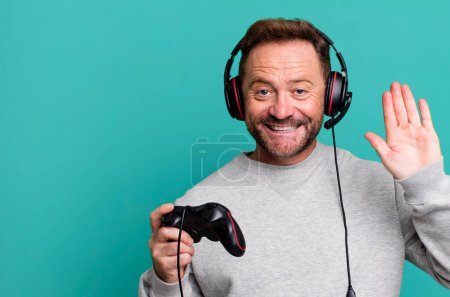 Photo for Middle age man smiling happily, waving hand, welcoming and greeting you. gamer concept with a control and headset - Royalty Free Image