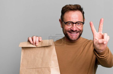 Foto de Hombre de mediana edad sonriendo y mirando feliz, haciendo gestos de victoria o paz. llevar bolsa de papel - Imagen libre de derechos
