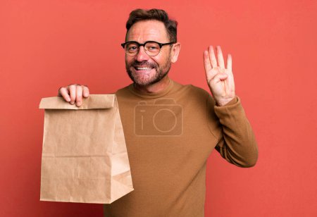 Téléchargez les photos : Homme d'âge moyen souriant et l'air amical, montrant le numéro quatre. emporter sac en papier - en image libre de droit