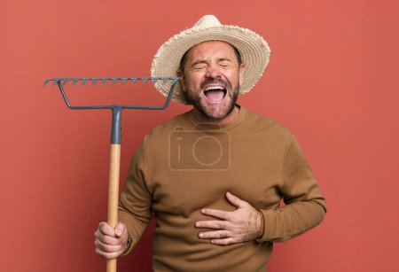 Téléchargez les photos : Homme d'âge moyen riant à haute voix à une blague hilarante. agriculteur avec un râteau - en image libre de droit