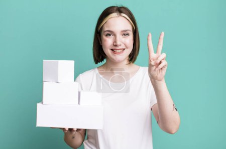 Téléchargez les photos : Jeune jolie femme souriante et heureuse, geste de victoire ou de paix. boîtes blanches vides - en image libre de droit