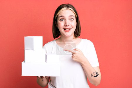 Photo for Young pretty woman looking excited and surprised pointing to the side. white blank boxes - Royalty Free Image