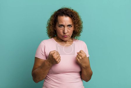 Téléchargez les photos : Femme hispanique d'âge moyen regardant confiant, en colère, fort et agressif, avec les poings prêts à se battre en position de boxe - en image libre de droit