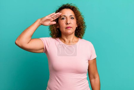 Téléchargez les photos : Femme hispanique du moyen âge saluant la caméra avec un salut militaire dans un acte d'honneur et de patriotisme, en montrant du respect - en image libre de droit