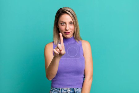 Photo for Blonde adult woman smiling and looking friendly, showing number one or first with hand forward, counting down - Royalty Free Image