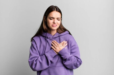 Photo for Pretty young adult woman looking sad, hurt and heartbroken, holding both hands close to heart, crying and feeling depressed - Royalty Free Image