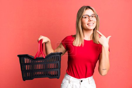 Foto de Young pretty woman smiling confidently pointing to own broad smile. empty shopping basket concept - Imagen libre de derechos