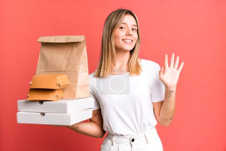 Photo for Young pretty woman smiling happily, waving hand, welcoming and greeting you. delivery and take away food concept - Royalty Free Image