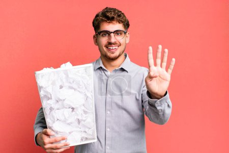 Téléchargez les photos : Jeune homme caucasien adulte souriant et regardant amical, montrant le numéro quatre avec un concept de poubelle de boules de papier - en image libre de droit