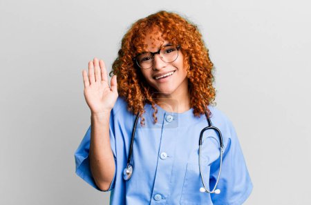 Téléchargez les photos : Cheveux roux jolie femme souriant joyeusement, agitant la main, vous accueillant et vous saluant. concept d'infirmière - en image libre de droit